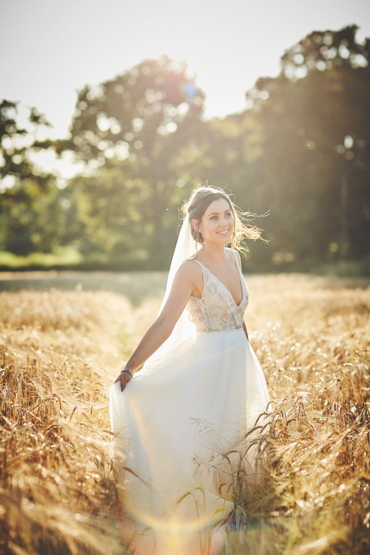 portrait of bride holding her wedding dress in field of corn or wheat at sunset at Upton Barn and Walled Garden in Devon